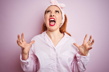 Young beautiful redhead woman wearing pajama standing over pink isolated background crazy and mad shouting and yelling with aggressive expression and arms raised. Frustration concept.