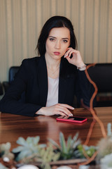 Young attractive emotional girl in business style clothes sitting at desk with phone in office or audience