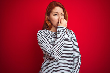 Young redhead woman wearing strapes navy shirt standing over red isolated background smelling...
