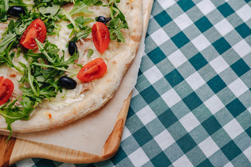 Neapolitan pizza with tuna, cheese, arugula, basil, tomatoes, olives, sprinkled with cheese on a wooden table on a tablecloth in a cell with a place for the text.