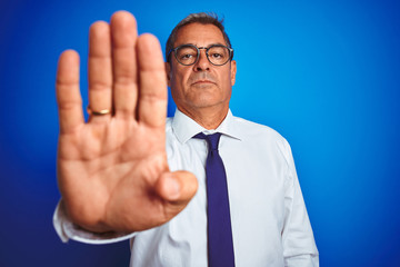 Handsome middle age businessman wearing glasses standing over isolated blue background with open hand doing stop sign with serious and confident expression, defense gesture