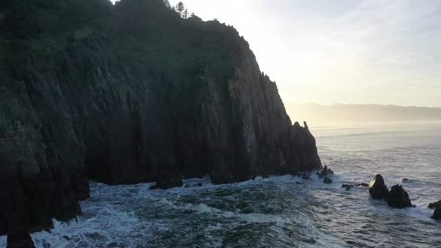 Aerial 4K video of rocky ocean cliff at sunrise. Hazy yellow dawn along Pacific Northwest coastline.