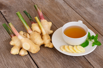 Fresh organic young ginger root, stem with slices and white cup of hot ginger tea with fresh green mint leaves isolated on wood table background . Natural herbal healthy drinks concept.Space for text.