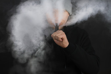 The man smoke an electronic cigarette on the dark background, Men in hoody vaping and releases a...