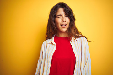 Young beautiful woman wearing red t-shirt and stripes shirt over yellow isolated background winking...