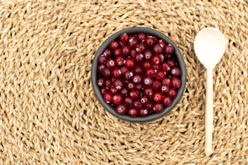 Top view at the bowl full of fresh cranberries standing at the table covered by wicker napkin background and wooden spoon laying at the side, rustic lifestyle