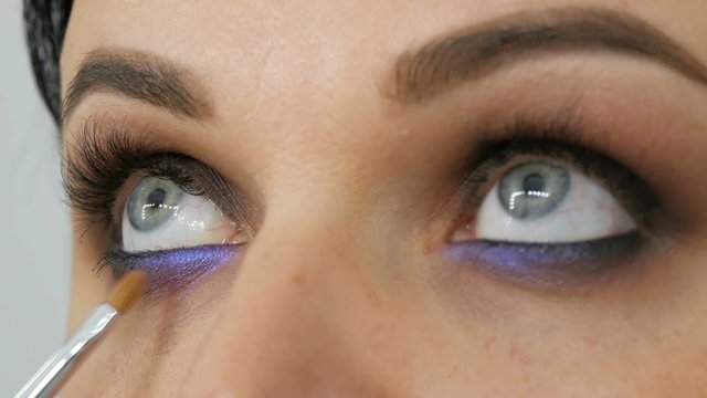 Beautiful blue eye to a young woman whom apply smoky eyes with the help of special brush Lilac and pearly smoky eyes eyeshadow, eyes and face of woman close up view. Professional high fashion.