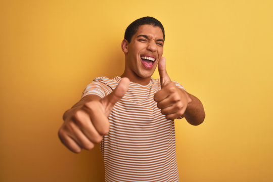 Young handsome arab man wearing striped t-shirt standing over isolated yellow background approving doing positive gesture with hand, thumbs up smiling and happy for success. Winner gesture.
