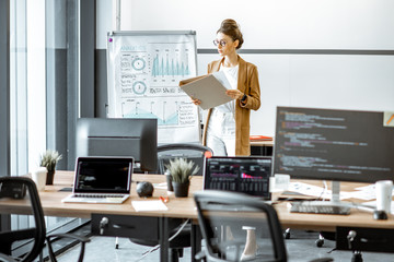 Fototapeta premium Young business woman preparing for a presentation, standing alone with documents near flipchart in the modern office or coworking space