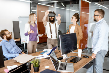 Group of diverse colleagues during small conference in the office, creative bearded man trying a new product with virtual reality goggles