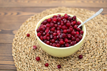 Healthy breakfast with frozen cranberries in big green bowl, metal spoon inside and spread berries at wooden table covered by wicker round napkin in the countryside