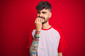 Young man with tattoo wearing striped t-shirt standing over isolated red background looking...