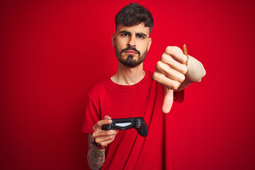 Young gamer man with tattoo playing video game standing over isolated red background with angry face, negative sign showing dislike with thumbs down, rejection concept