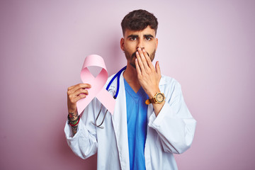 Young doctor man with tattoo holding cancer ribbon standing over isolated pink background cover mouth with hand shocked with shame for mistake, expression of fear, scared in silence, secret concept