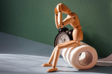 Wooden figure sit on a roll of toilet paper near alarm clock. Concept of the problem with digestion.