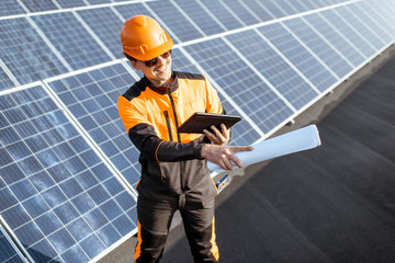 Portrait of an engineer in protective workwear carrying out service of solar panels with digital tablet and blueprintson a photovoltaic rooftop plant