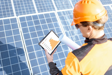 Workman examining genaration of solar power plant, holding digital tablet with a chart of...