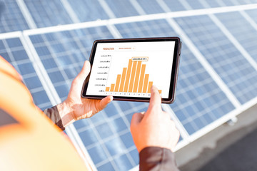 Workman examining genaration of solar power plant, holding digital tablet with a chart of...