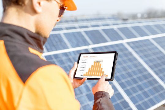 Workman Examining Genaration Of Solar Power Plant, Holding Digital Tablet With A Chart Of Electricity Production. Concept Of Online Monitoring Of The Electric Station