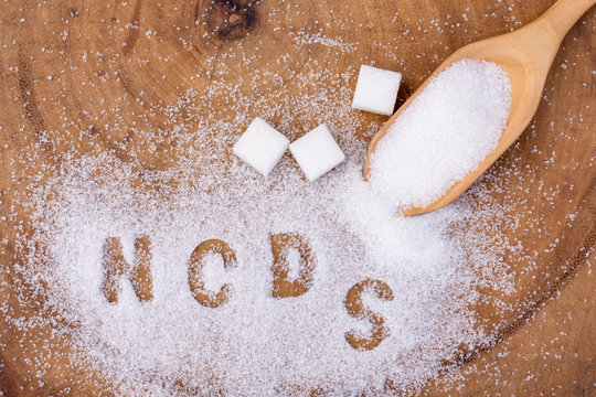 Closeup white granulated sand sugar in wooden spoon and words"NCDs"  letter written in sugar grains on wood table  background.Unhealthy diet ,awareness and stop NCD  concept. Top view. Flat lay.