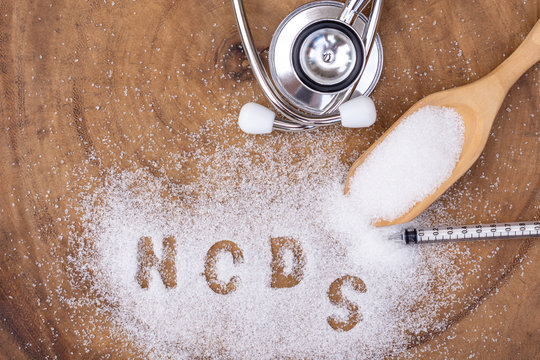 Closeup white granulated sand sugar in wooden spoon and words"NCDs"  letter written in sugar grains with medical stethoscope on wood table  background Top view. Flat lay.
