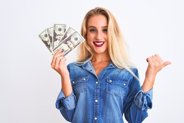 Young beautiful blond woman holding dollars standing over isolated white background pointing and showing with thumb up to the side with happy face smiling