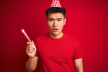 Asian chinese man on birthday celebration wearing funny hat over isolated red background scared in...