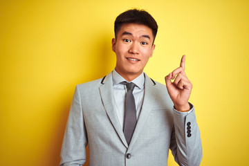 Asian chinese businessman wearing suit and tie standing over isolated yellow background pointing finger up with successful idea. Exited and happy. Number one.