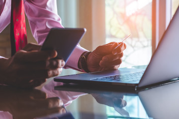 Casual businessman hand holding credit card and using mobile smart phone for online shopping and payment, work on laptop computer on the wooden table.