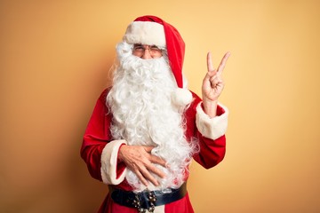 Fototapeta na wymiar Middle age handsome man wearing Santa costume standing over isolated yellow background smiling with happy face winking at the camera doing victory sign. Number two.
