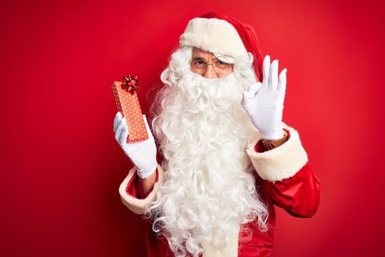 Middle age man wearing Santa Claus costume holding gift over isolated red background doing ok sign with fingers, excellent symbol