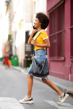 Full Length Side Of Happy Young Black Woman Walking And Talking With Cellphone On Street