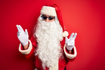 Middle age man wearing Santa Claus costume and sunglasses over isolated red background with serious expression on face. Simple and natural looking at the camera.