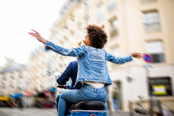 behind of young african american woman sitting on back of scooter driving through city streets with...