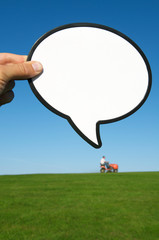 Blank speech bubble sign with copy space above a distant lawn mower tractor on the green grass horizon under sunny blue sky
