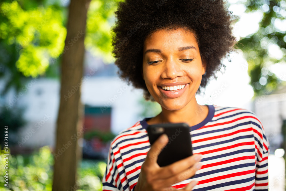 Wall mural Close up beautiful young black woman with afro hair smiling at mobile phone in park