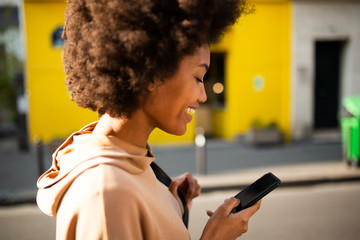 Side of beautiful young smiling black woman with afro hairstyle walking with cellphone in city