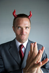 Cunning evil businessman with red devil horns and suit rubbing his hands together in anticipation with a conniving expression on his face