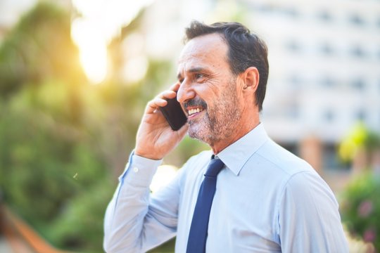 Middle age handsome businessman standing on the street talking on the smartphone smiling