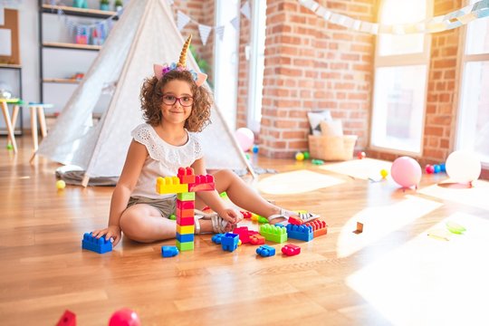 Beautiful toddler wearing glasses and unicorn diadem sitting playing with building blocks smiling at kindergarten