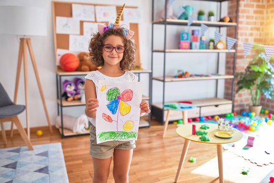 Beautiful toddler wearing glasses and unicorn diadem standing holding draw smiling at kindergarten