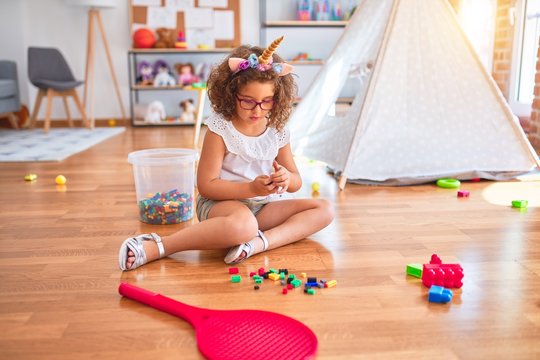 Beautiful toddler wearing glasses and unicorn diadem sitting on the floor playing with building blocks at kindergarten
