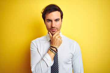 Young handsome businessman wearing elegant shirt and tie over isolated yellow background looking confident at the camera with smile with crossed arms and hand raised on chin. Thinking positive.