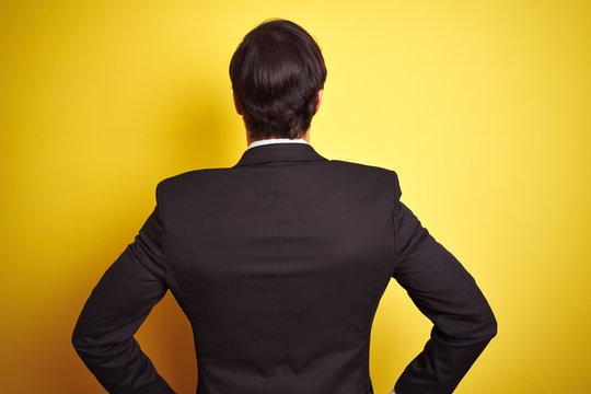 Young handsome businessman wearing suit and tie standing over isolated yellow background standing backwards looking away with arms on body