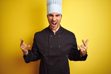 Young chef man wearing uniform and hat standing over isolated yellow background shouting with crazy...