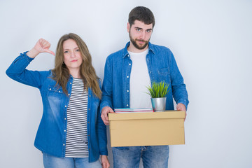 Young couple holding cardboard box moving to new house over white isolated background with angry face, negative sign showing dislike with thumbs down, rejection concept