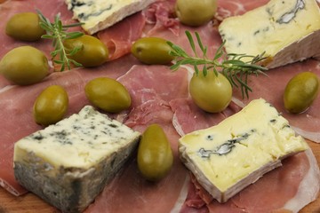 Sliced jamon, green olives, pieces of blue cheese and sprig of lavender laid out on a wooden board, background