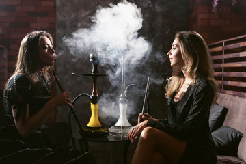 Girls party in hookah lounge. Group of two young women in backlight smoking shisha in cafe or bar,...