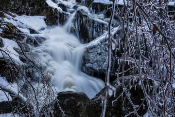 Brook of water in winter nature. Cold stream water flows between snowed stones. Iced branches in front of waterfall.
