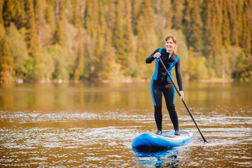 Girl in thermo clothing rowing oar on sup board blue lake water paddleboard background of forest